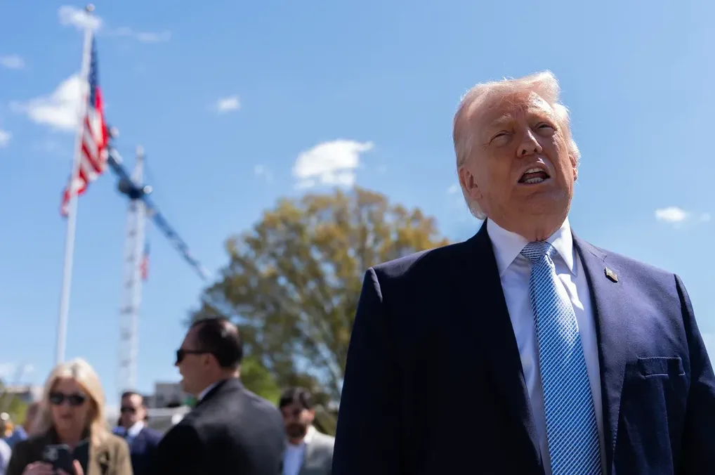 Donald Trump u tamnom odijelu i plavoj kravati govori na otvorenom, s američkom zastavom i dizalicom u pozadini.
