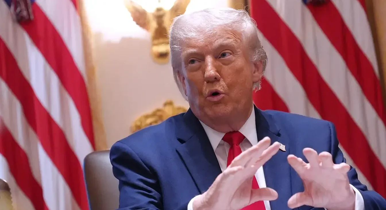 Donald Trump u plavom odijelu i crvenoj kravati, sa podignutim rukama i izrazom na licu, ispred američkih zastava.