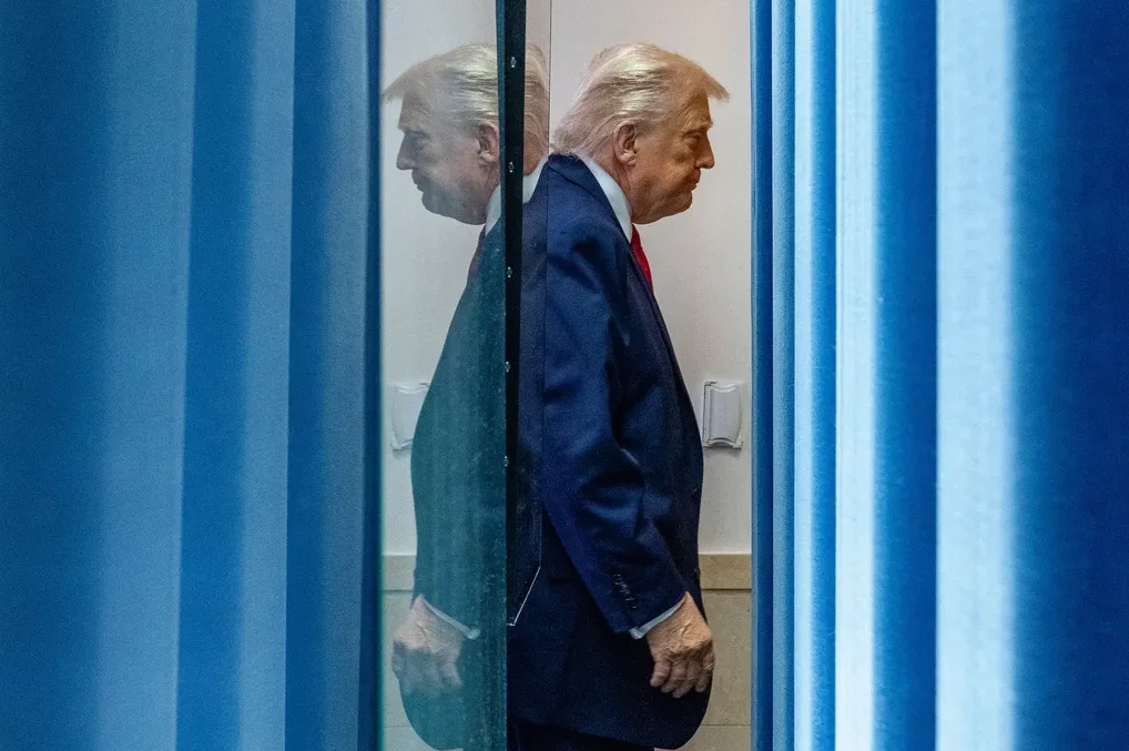 Donald Trump gleda u stranu, sa svojom refleksijom u staklenoj pregradi iza plavih vertikalnih zavjesa.