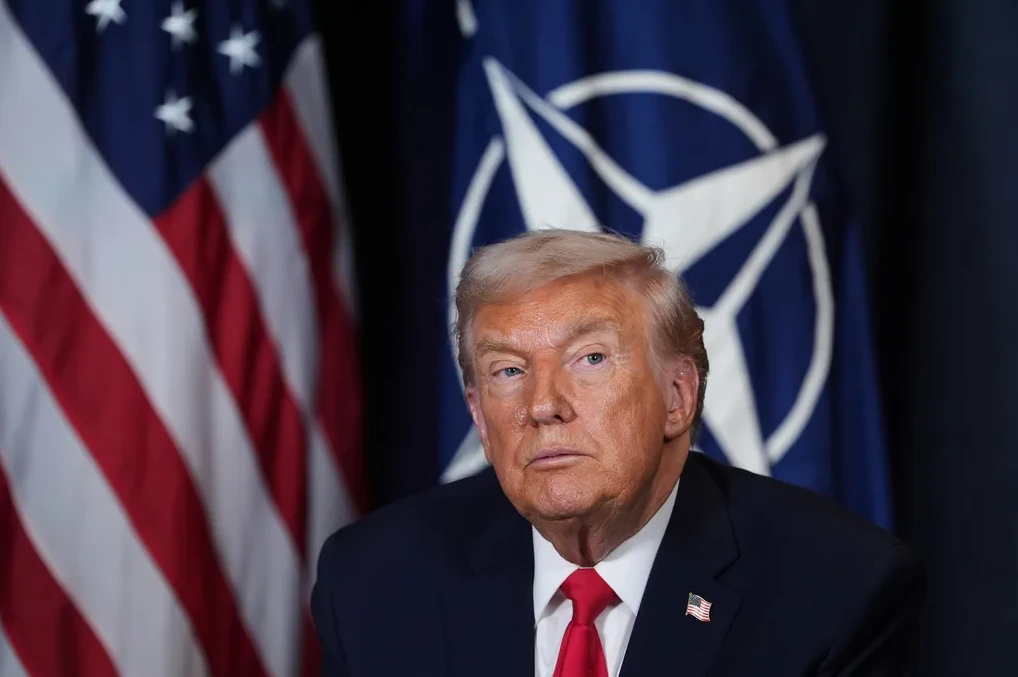 Donald Trump u tamnom odijelu i crvenoj kravati ispred zastave SAD-a i NATO-a