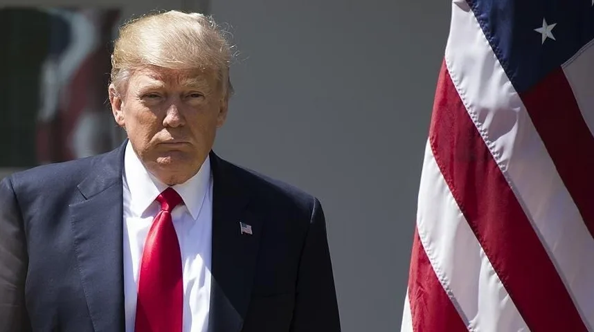 Donald Trump u tamnom odijelu i crvenoj kravati stoji pored američke zastave.