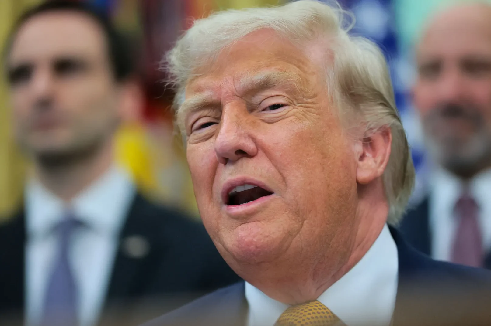 Donald Trump govori u Bijeloj kući, sa dva nejasna muškarca u pozadini.