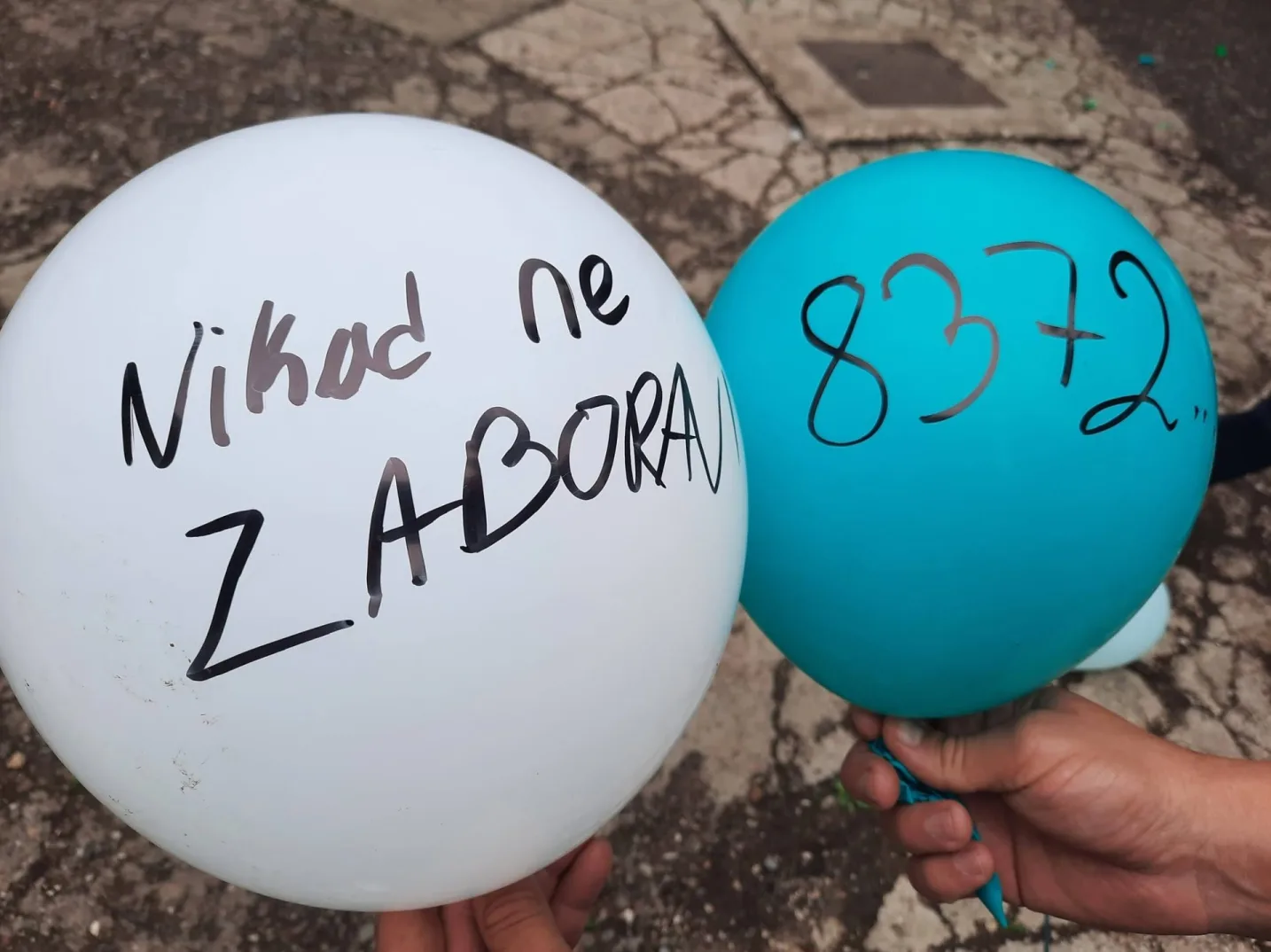 Dva balona, jedan bijeli i jedan plavi, sa ispisanim porukama. Na bijelom balonu piše "Nikad ne ZABORAV", a na plavom balonu je broj "8372".