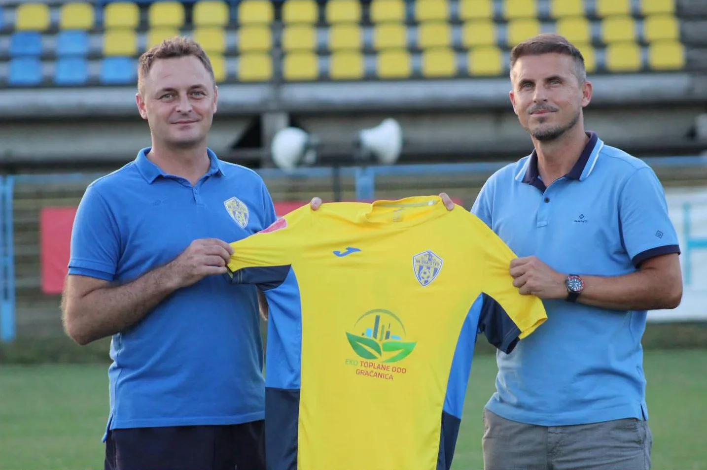 Dva muškarca u plavim majicama drže žutu fudbalsku majicu sa grbom NK Bratstvo i logom sponzora Eko Toplane doo Gračanica.