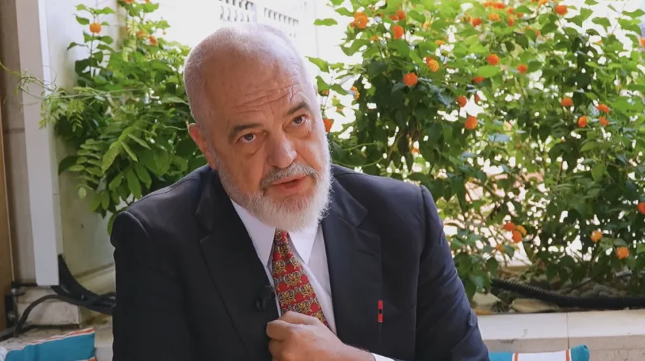 Edi Rama, premijer Albanije, sjedi u odijelu sa crvenom kravatom i bijelom košuljom, sa bijelom bradom i kratkom sijedom kosom. Pozadina je zelena sa narandžastim cvjetovima.