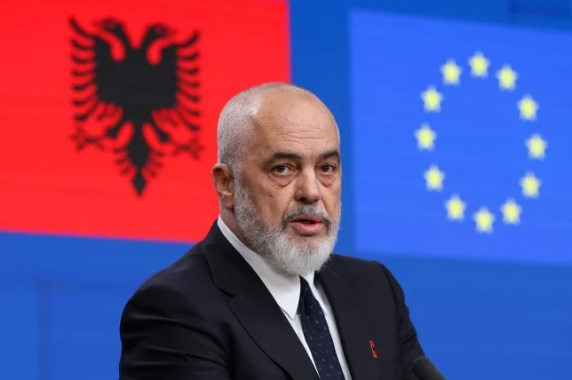 Edi Rama, premijer Albanije, u crnom odijelu, bijeloj košulji i tamnoj kravati, govori pred zastavom Albanije (crvena sa crnim dvoglavim orlom) i zastavom Evropske unije (plava sa krugom žutih zvjezdica).