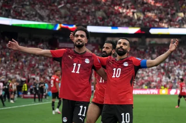 Fudbaleri Egipta u crvenim dresovima sa brojevima 10 i 11 slave postignut gol, sa publikom u pozadini.