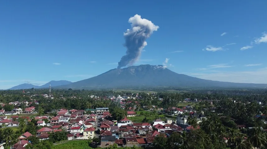 Dim iz vulkana diže se visoko u nebo iznad planine, sa naseljem u prvom planu.