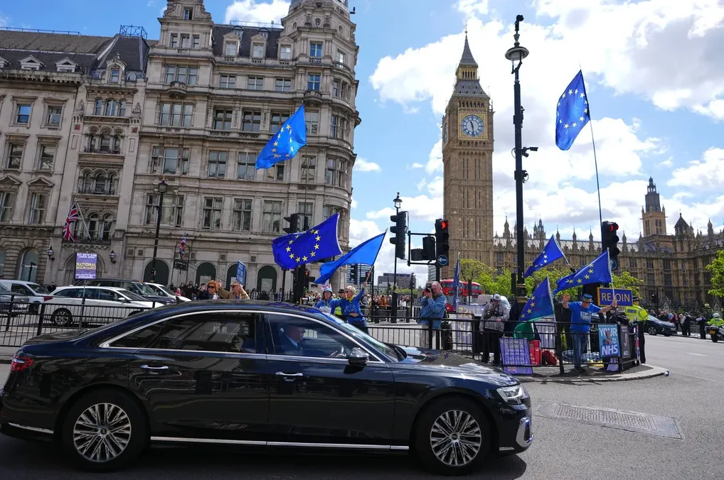 Crni luksuzni automobil prolazi pored grupe ljudi koji mašu zastavama Evropske unije ispred zgrade Parlamenta i Big Bena u Londonu.