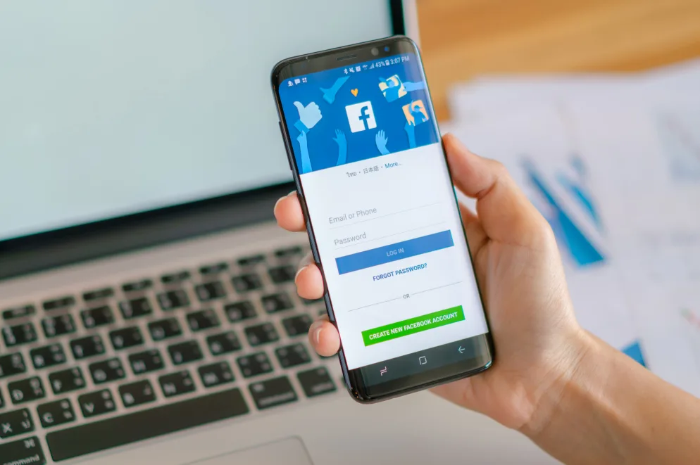 Ruka drži pametni telefon s Facebookovim ekranom za prijavu, pored laptopa.