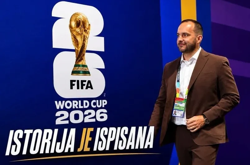 Čovjek u smeđem odijelu stoji pored postera FIFA Svjetskog prvenstva 2026.