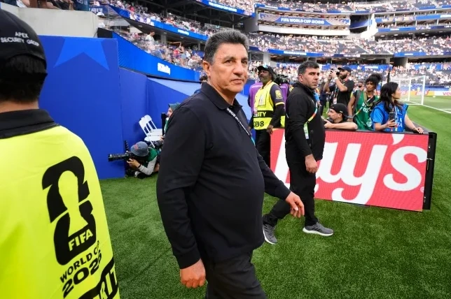 Muškarac u crnoj majici sa oznakom "FIFA WORLD CUP 2022" na kragni, u pratnji službenika obezbeđenja na stadionu.