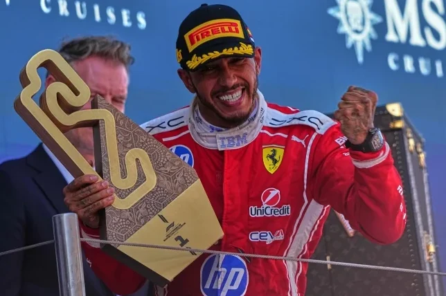 Vozač Formule 1 u crvenom odijelu slavi sa podignutom pesnicom i drži trofej u obliku staze.