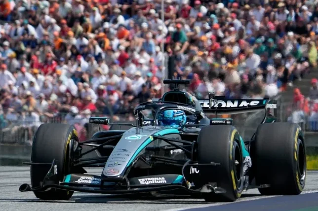 Mercedes F1 bolid sa vozačem u plavom odijelu, u punoj brzini na trkačkoj stazi, sa gledaocima u pozadini.