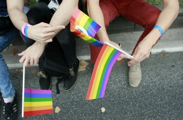 Dvije osobe sjede na ivici pločnika i drže male duge zastave u rukama, simbolizujući podršku LGBT zajednici.
