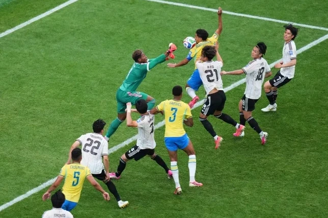 Fudbaleri u bijelim dresovima sa crnim prugama i brazilski fudbaleri u žutim dresovima takmiče se za loptu na zelenom terenu tokom utakmice.