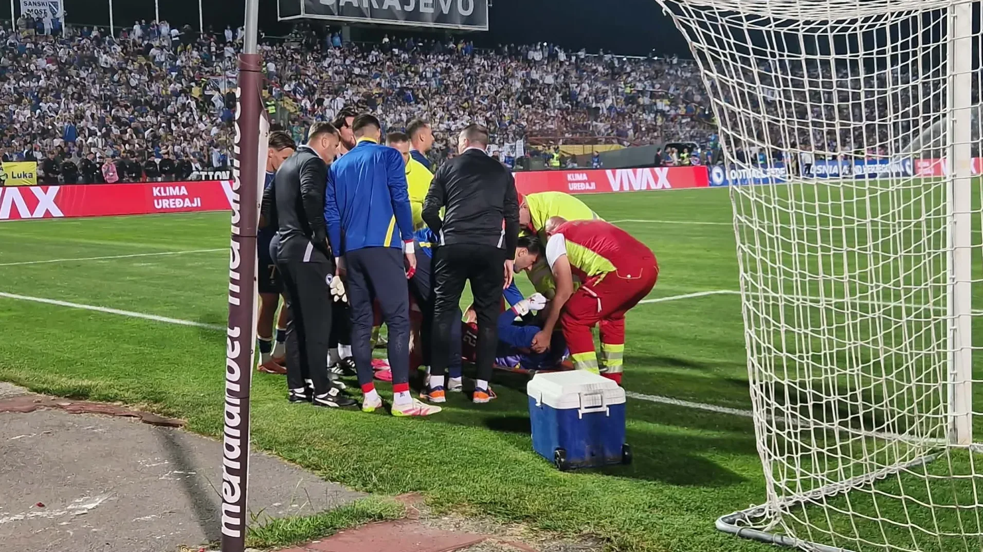 Fudbaler leži na travi, medicinska ekipa mu pruža pomoć na terenu, iza njega su saigrači i publika na stadionu.