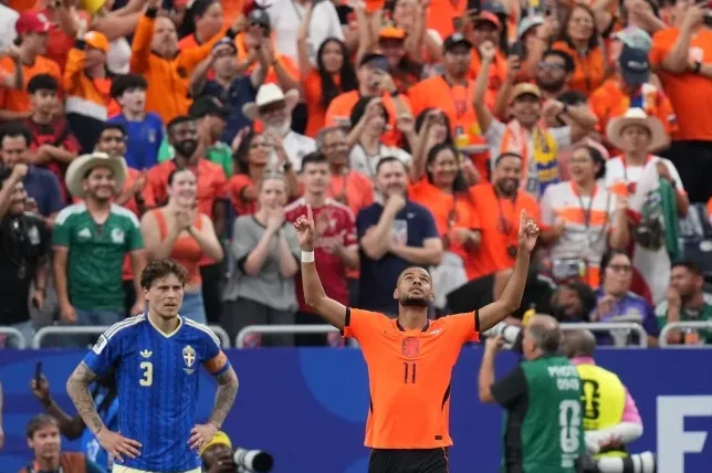Fudbaler u narandžastoj majici sa brojem 11 slavi gol, dok njegov saigrač u plavoj majici sa brojem 3 stoji pored njega razočaranog izraza lica, a iza njih se vidi navijačka publika na stadionu.