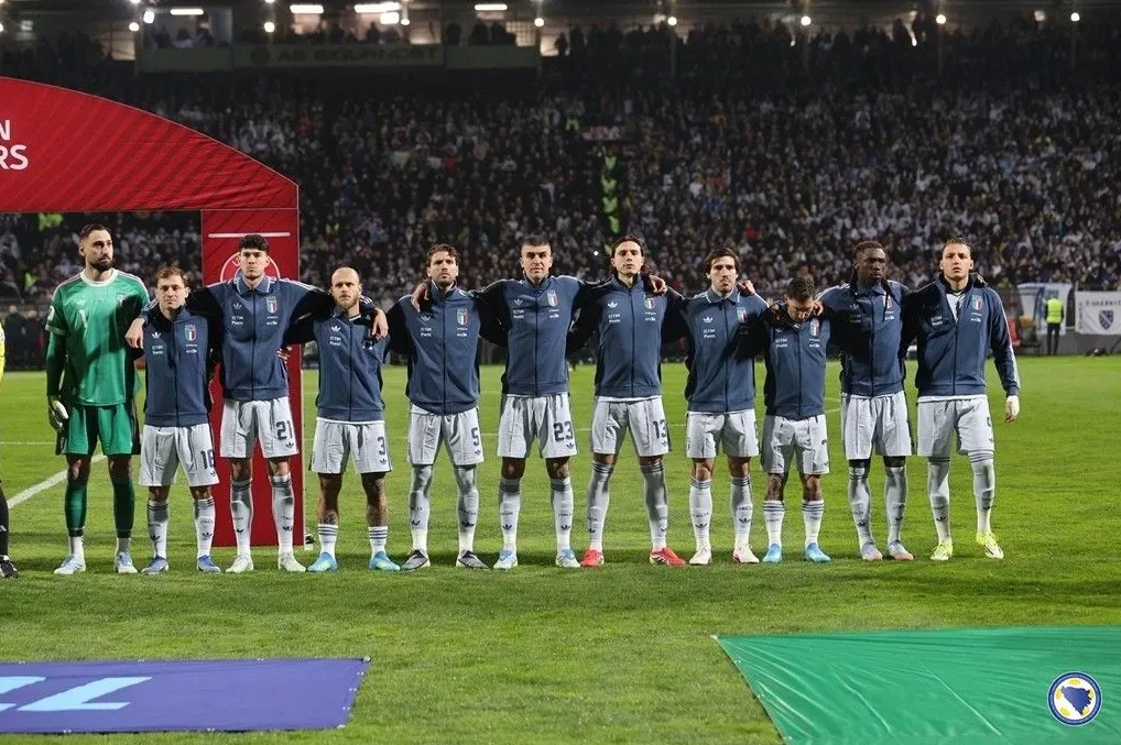 Fudbalska reprezentacija Italije postrojena na terenu prije utakmice, sa navijačima na tribinama u pozadini.