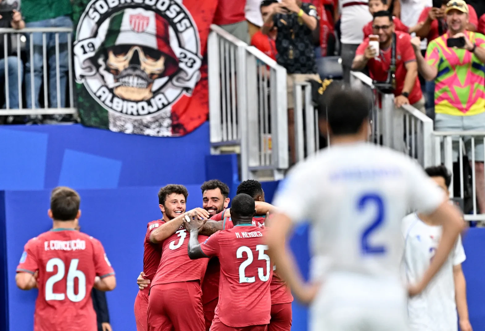 Fudbaleri u crvenim dresovima grle se slaveći gol, sa navijačima u pozadini.
