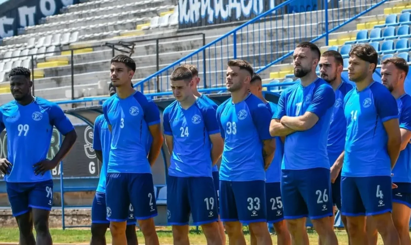 Grupa fudbalera u plavim dresovima stoji u redu na stadionu.