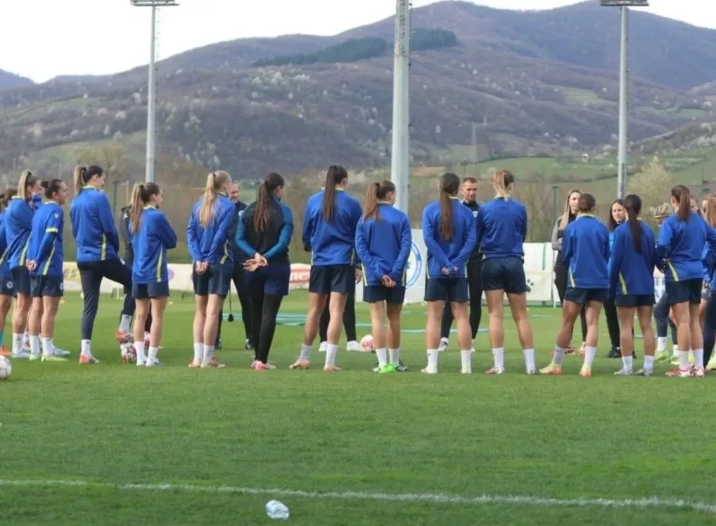 Ženski fudbalski tim u plavim dresovima na treningu, okupljeni na zelenom terenu sa brdima u pozadini.