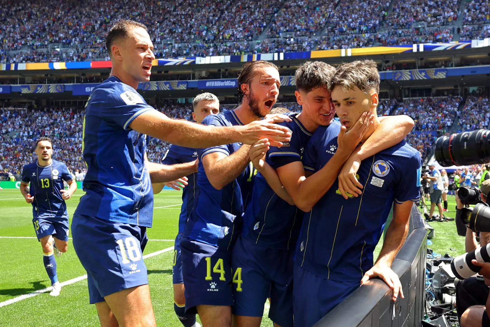 Fudbaleri u plavim dresovima grle jedni druge na terenu, slaveći gol ili pobjedu, dok ih publika na tribinama bodri.