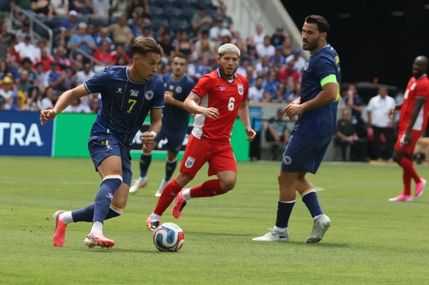 Fudbaleri u tamnoplavim i crvenim dresovima takmiče se na zelenom terenu stadiona tokom utakmice.