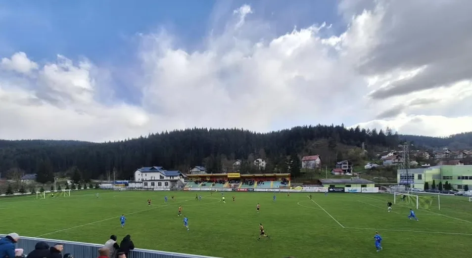 Fudbalski teren sa igračima u plavim i crvenim dresovima, okružen tribinama i šumovitim brdima pod oblačnim nebom.