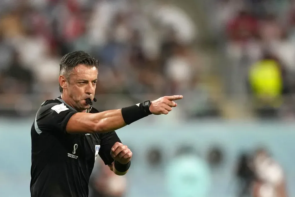 Fudbalski sudija sa zviždaljkom u ustima, pokazujući kažiprstom u pravcu terena, noseći crni dres sa oznakama FIFA Svjetskog prvenstva u Kataru 2022.