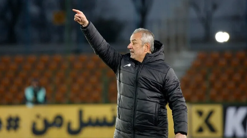 Fudbalski trener u crnoj jakni sa kapuljačom, sijede kose, podignute desne ruke i ispruženog kažiprsta usmjerenog ulijevo, na fudbalskom stadionu, sa tribinama i reklamama u pozadini.