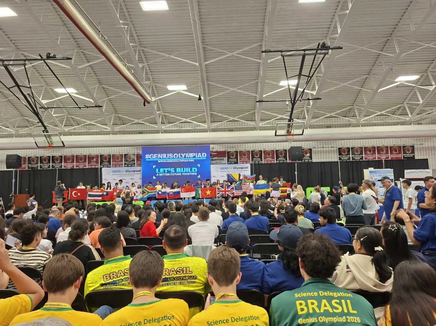 Učesnici Genius Olympiad takmičenja u velikoj dvorani, ispred bine sa zastavama zemalja i velikim ekranom na kojem piše "#GENIUSOLYMPIAD LET'S BUILD A BETTER FUTURE TOGETHER"