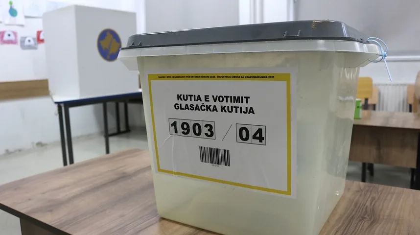 Providna glasačka kutija sa sivim poklopcem, sa žutom naljepnicom na kojoj piše "Kutia e votimit / Glasačka kutija" i brojevima "1903 / 04". U pozadini se vidi biračko mjesto sa kutijom za glasanje sa grbom Kosova.