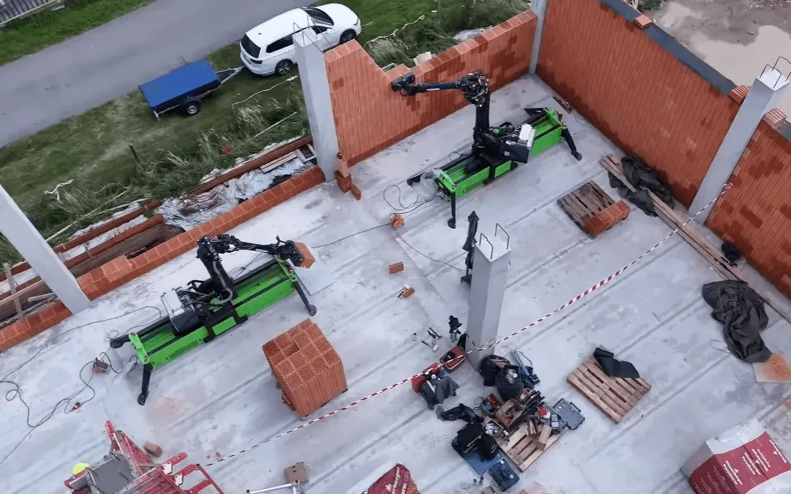 Robotski zidari postavljaju cigle na gradilištu, u blizini se nalazi bijeli automobil s prikolicom.