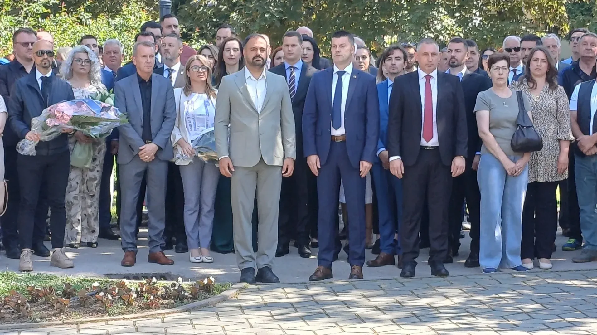 Grupa muškaraca i žena u svečanoj odjeći, neki drže cvijeće.