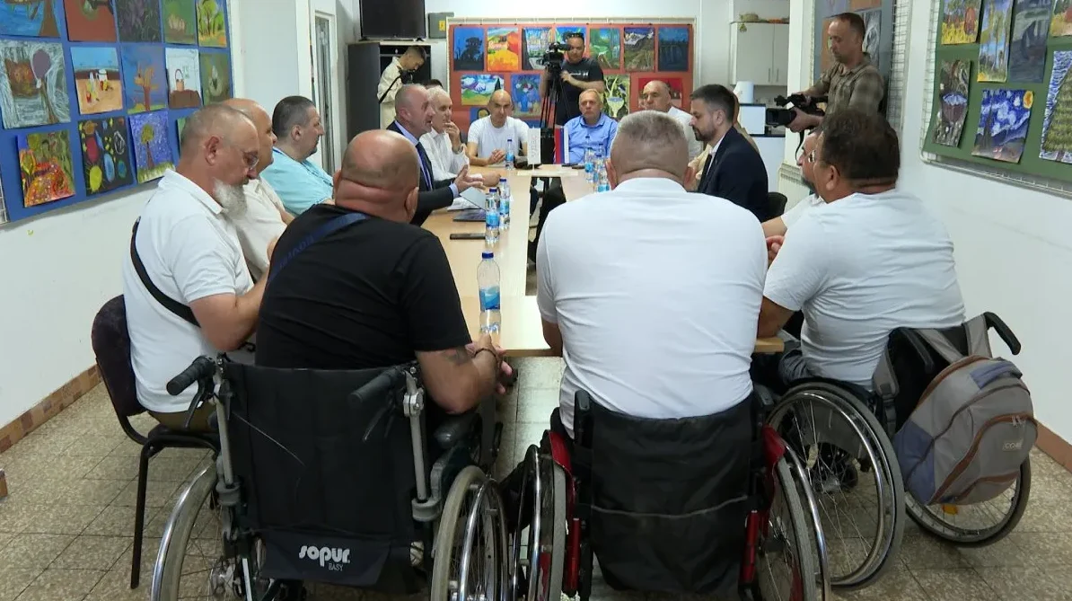 Grupa muškaraca, od kojih neki sjede u invalidskim kolicima, okupljeni su oko stola na sastanku. Na zidovima prostorije vise dječiji crteži.