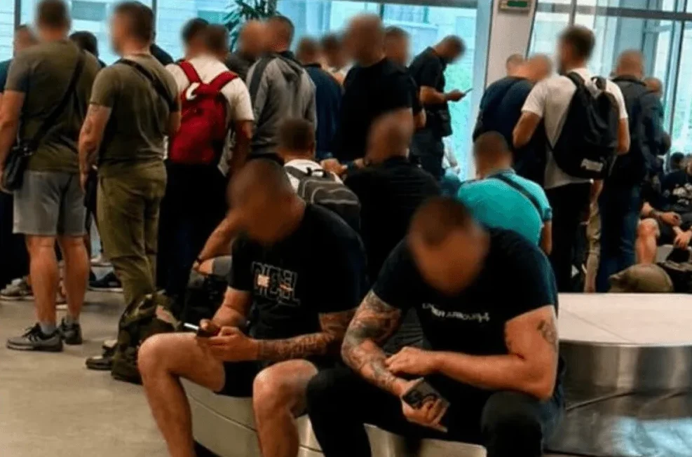 Grupa muškaraca, od kojih su neki sjedili, a neki stajali, čekaju u zatvorenom prostoru.
