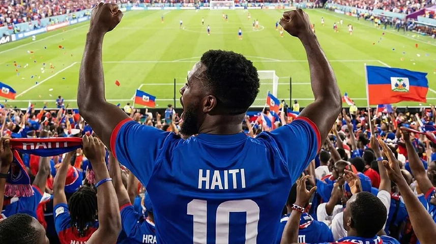 Fudbalski navijač Haitija, okrenut leđima kameri, podiže ruke u zrak slaveći sa zastavom Haitija i drugim navijačima.