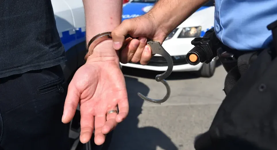Policajac stavlja lisice uhapšeniku na ruku, sa službenim automobilom u pozadini.