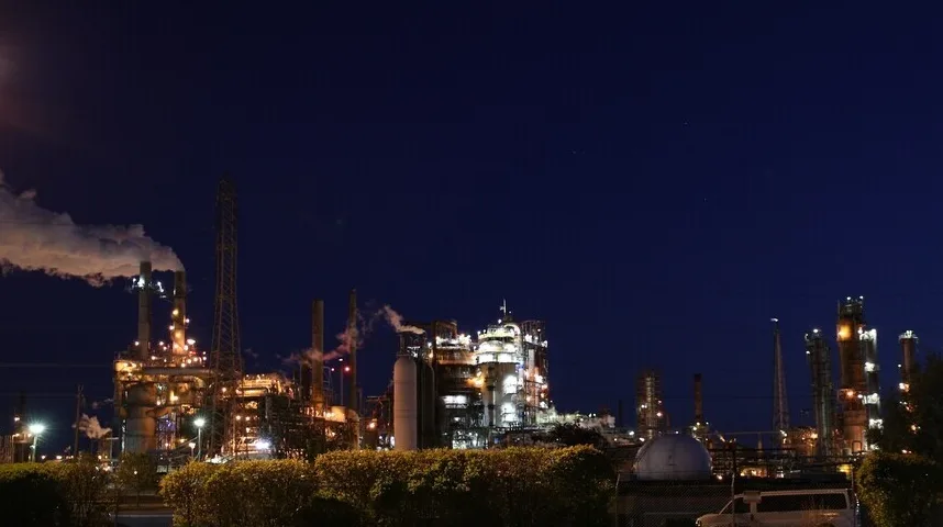 Industrijska postrojenja osvijetljena reflektorima pod tamnim noćnim nebom, sa parom koja izlazi iz dimnjaka.