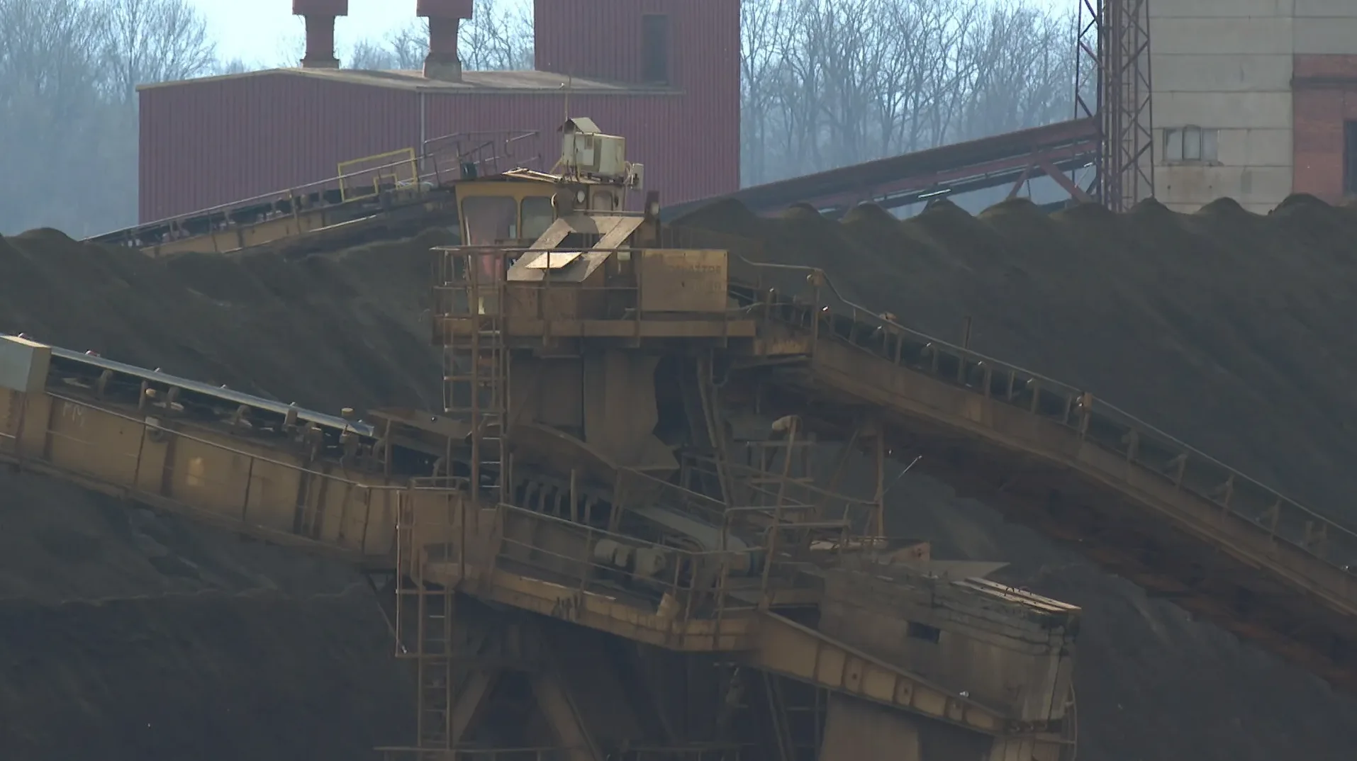 Detaljan pogled na industrijski manipulator postavljen iznad pokretne trake u rudniku, uz brda crnog materijala u pozadini.