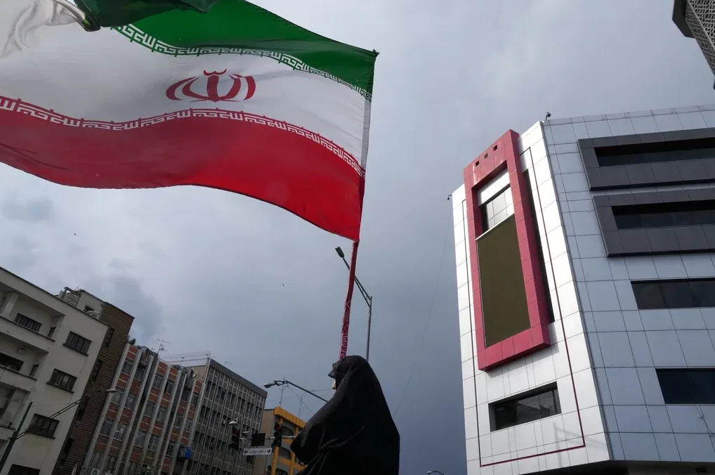 Iranska zastava vijori se na vjetru ispred modernih zgrada i oblačnog neba.