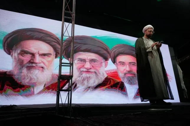 Veliki ekran prikazuje portrete dvojice iranskih vjerskih vođa, a desno stoji muškarac u vjerskoj odjeći gledajući u telefon.