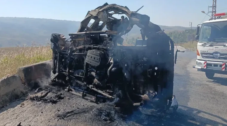 Potpuno izgorjelo vozilo prevrnuto na putu, pored njega se vidi vatrogasno vozilo.