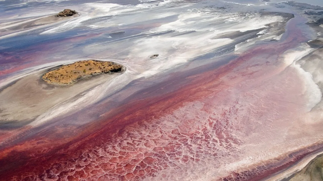 Gornji pogled na jezero sa intenzivnom roze bojom vode, koje formira karakteristične šare i forme na površini. U vodi se nazire malo, suvo ostrvo obraslo niskom vegetacijom.