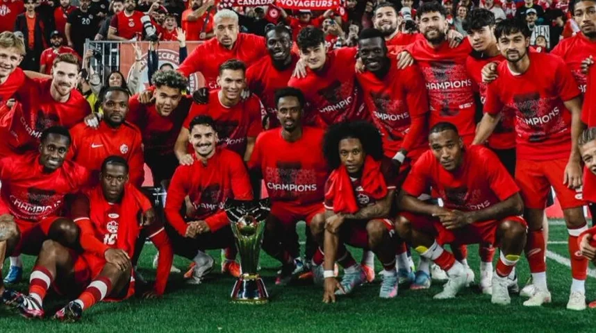 Kanadski nogometni tim u crvenim dresovima sa natpisom "Šampioni", slaveći pobjedu sa podignutim trofejem.