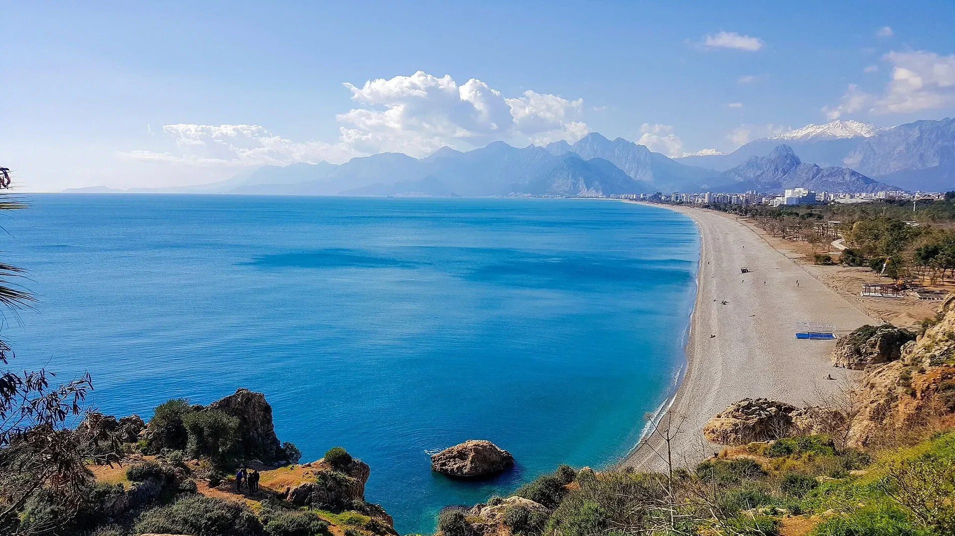 Pogled na šljunkovitu plažu Konyaalti u Antaliji, Turska, s plavim Mediteranskim morem i planinama u pozadini.