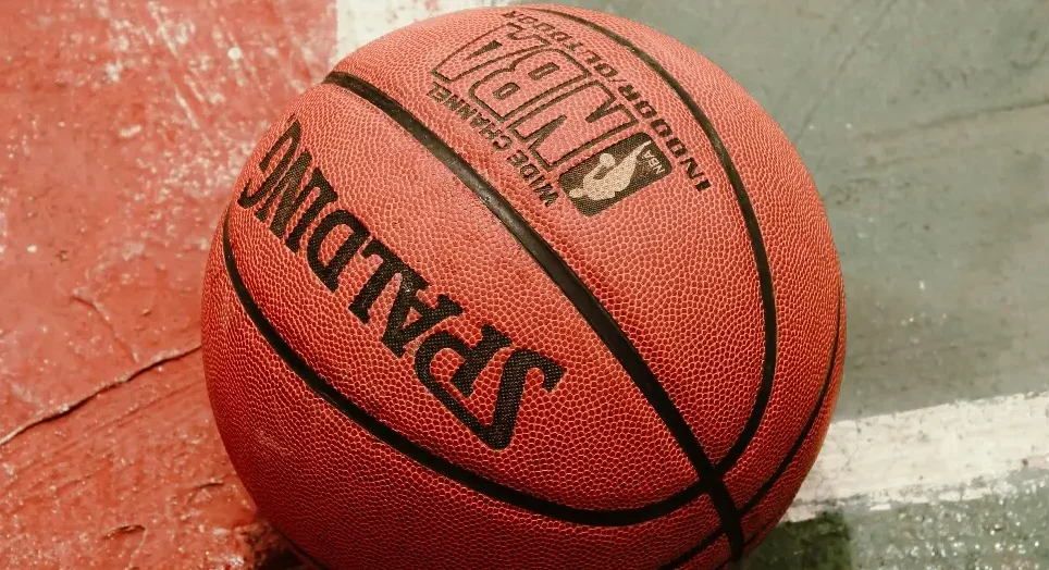 Detaljan pogled na crvenu Spalding košarkašku loptu sa crnim linijama, sa logom NBA i natpisom "INDOOR/OUTDOOR" i "WIDE CHANNEL". Lopta leži na podlozi koja podsjeća na košarkaški teren sa crvenim i sivim dijelovima.