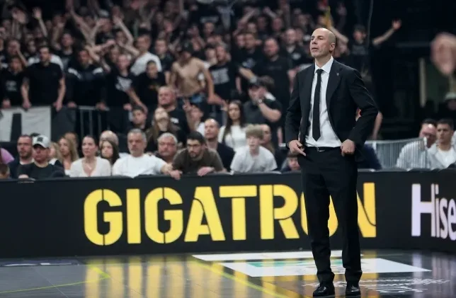 Košarkaški trener u crnom odijelu stoji na terenu, gleda u publiku.