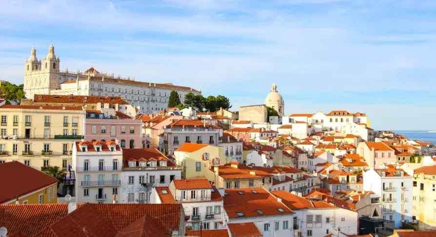 Panoramski pogled na grad Lisabon s mnoštvom kuća sa crvenim krovovima i bijelim fasadama, te impozantnom crkvom u pozadini pod vedrim plavim nebom.